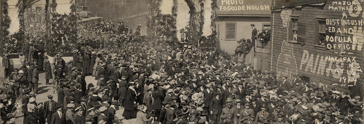 Soldiers participating in a parade following their return to Canada following the First World War. A large crowd has gathered to welcome their arrival at a wharf in Halifax. The soldiers appear to be carrying Major Matheson on their shoulders in the bottom right corner of the photograph. A handwritten note on the photo credits it to one "F.G. Goodenough." The ship from which the soldiers are disembarking is partially visible in the background.