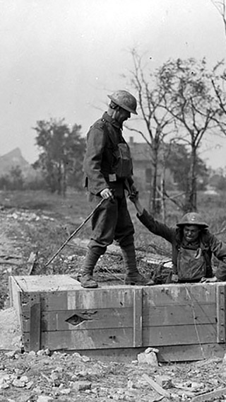 On voit dans l’image en noir et blanc deux soldats canadiens qui émergent d’un tunnel allemand souterrain en France. Un des soldats est debout donnant un coup de main à l’autre soldat qui est en train de sortir du tunnel. On voit des bâtiments bombardés en arrière-plan.